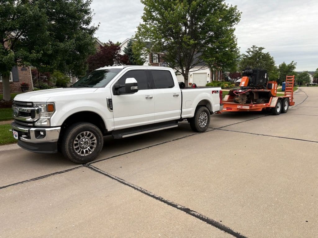 White Ford truck towing an orange trailer with equipment parked on a residential street.